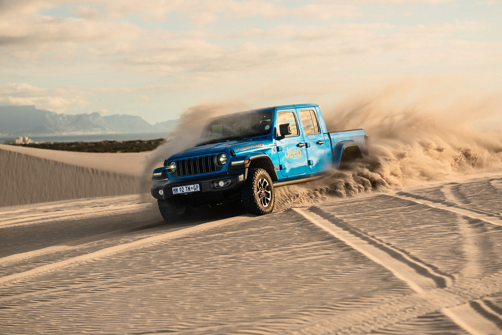 Blue Jeep Gladiator pickup drifting across desert sand dunes, kicking up dust with mountains in the background