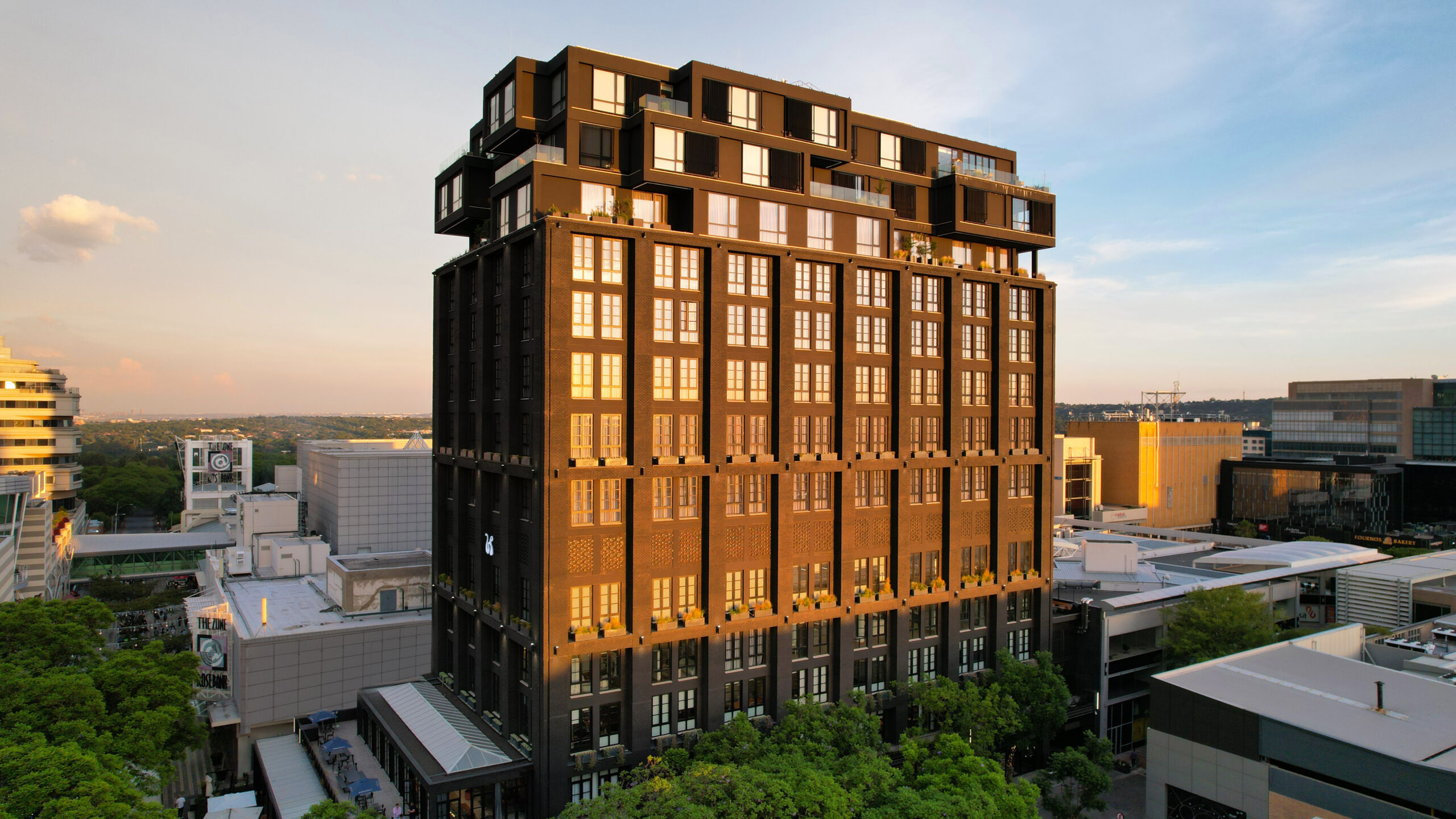 Exterior of The Bank in Rosebank, Johannesburg, showing the restored heritage building with its dark façade, framed by city streets, trees, and people moving through the precinct. The space reflects Rosebank’s urban lifestyle, where historic architecture meets contemporary design, hospitality, and everyday city life.