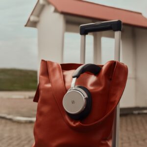 Bang & Olufsen Beoplay H100 headphones in Hourglass Sand, Beo Grace earphones in Honey Tone and a Beosound A1 speaker in Eucalyptus Green are arranged beside travel essentials.