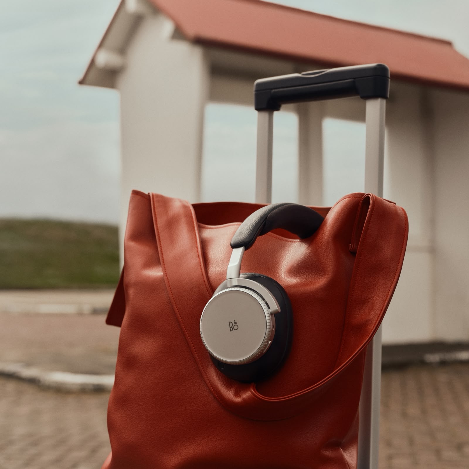 Bang & Olufsen Beoplay H100 headphones in Hourglass Sand, Beo Grace earphones in Honey Tone and a Beosound A1 speaker in Eucalyptus Green are arranged beside travel essentials.
